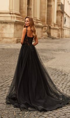 A woman in the La Femme 33499 long prom dress, featuring a black sheer floor-length skirt and fitted lace bodice with delicate details, stands on a cobblestone street, looking over her shoulder.