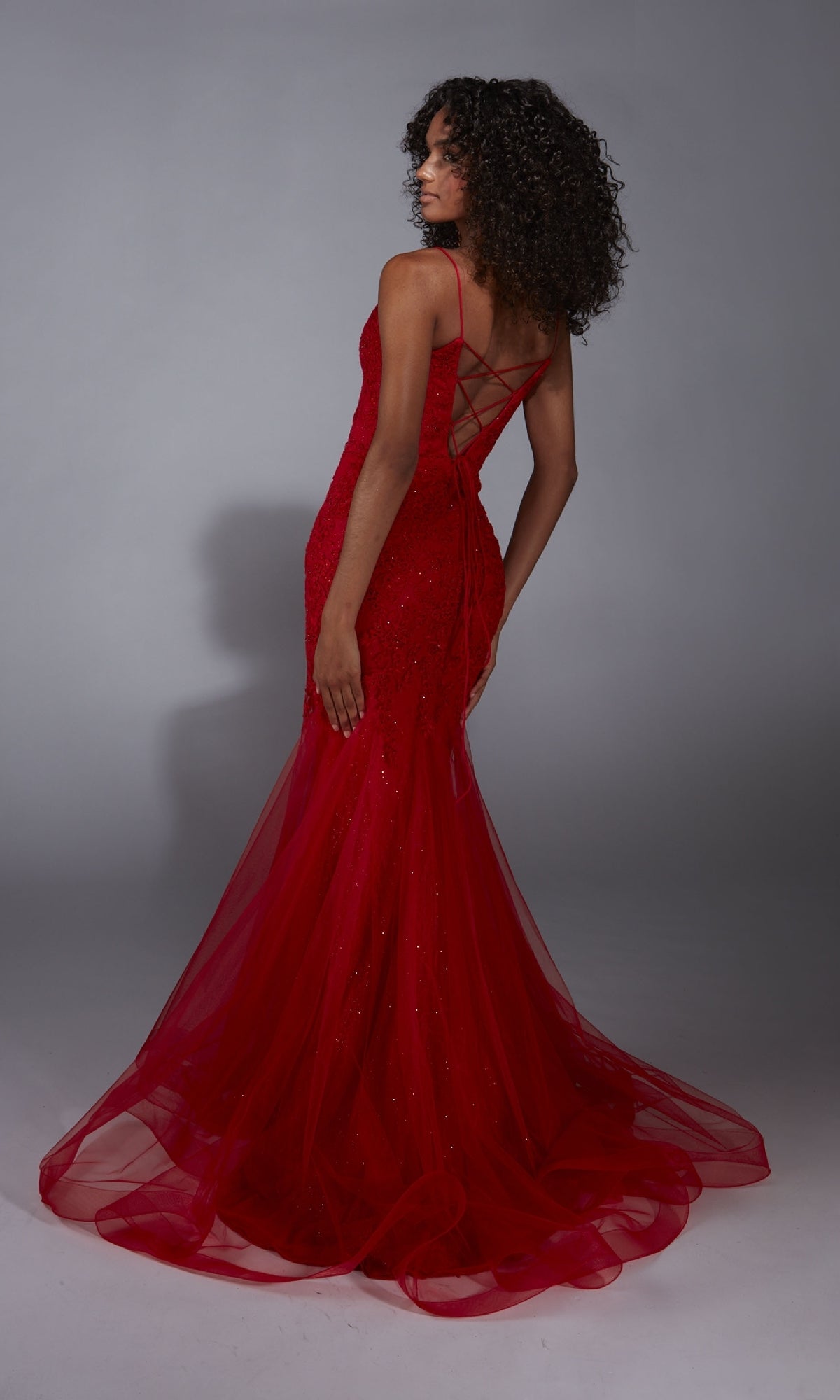 A woman stands facing away in a studio, wearing the Aerie Couture 334—a fitted, floor-length red mermaid prom dress featuring thin straps and a lace-up back.