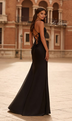 A woman stands outdoors on a stone plaza in front of a building, wearing the La Femme 33505 long prom dress—a black, backless evening gown with a plunging neckline and lace-up detailing.