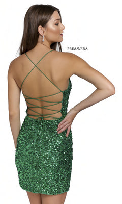 A woman stands sideways and faces away in a Short Sequin Lace-Up Party Dress 3351D, featuring a green sequin design and open lace-up back. She is set against a plain white background with "PRIMAVERA" text visible.
