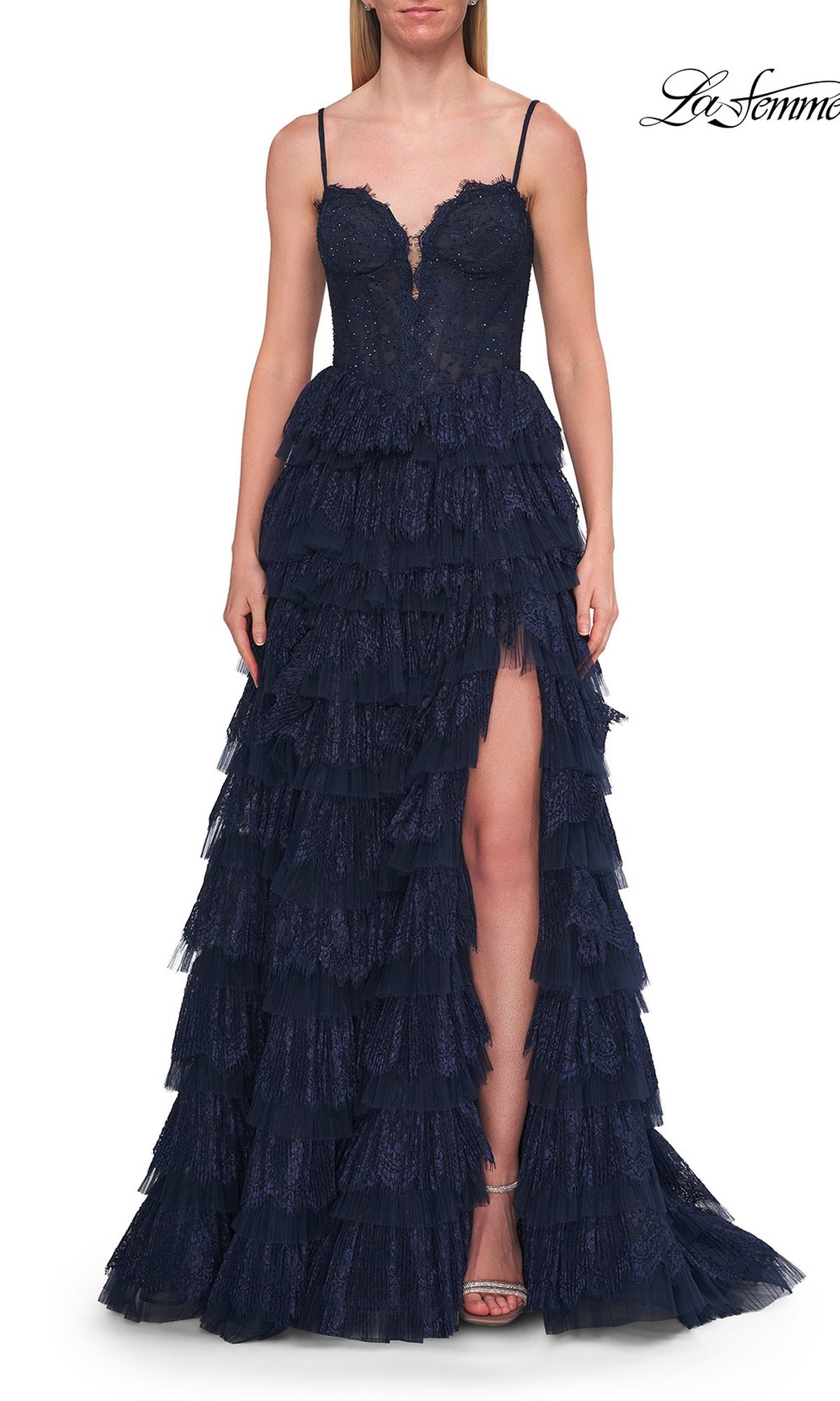 A woman wearing the La Femme 33525 long prom dress in marine blue, featuring a lace tiered skirt, corset bodice, spaghetti straps, and a thigh-high slit, stands against a white background.