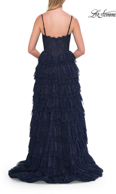 A woman is wearing the Long Prom Dress: La Femme 33525—a navy blue, tiered lace ball gown with thin straps—photographed from the back to highlight its ruffled layers and fitted corset bodice, making it an ideal choice for an unforgettable prom look.