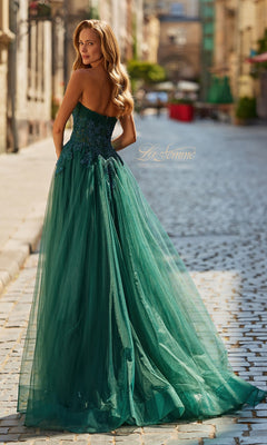 A woman stands on a cobblestone street, looking over her shoulder, wearing the La Femme 33528 long prom dress featuring a strapless sheer corset bodice and a floor-length emerald green skirt.