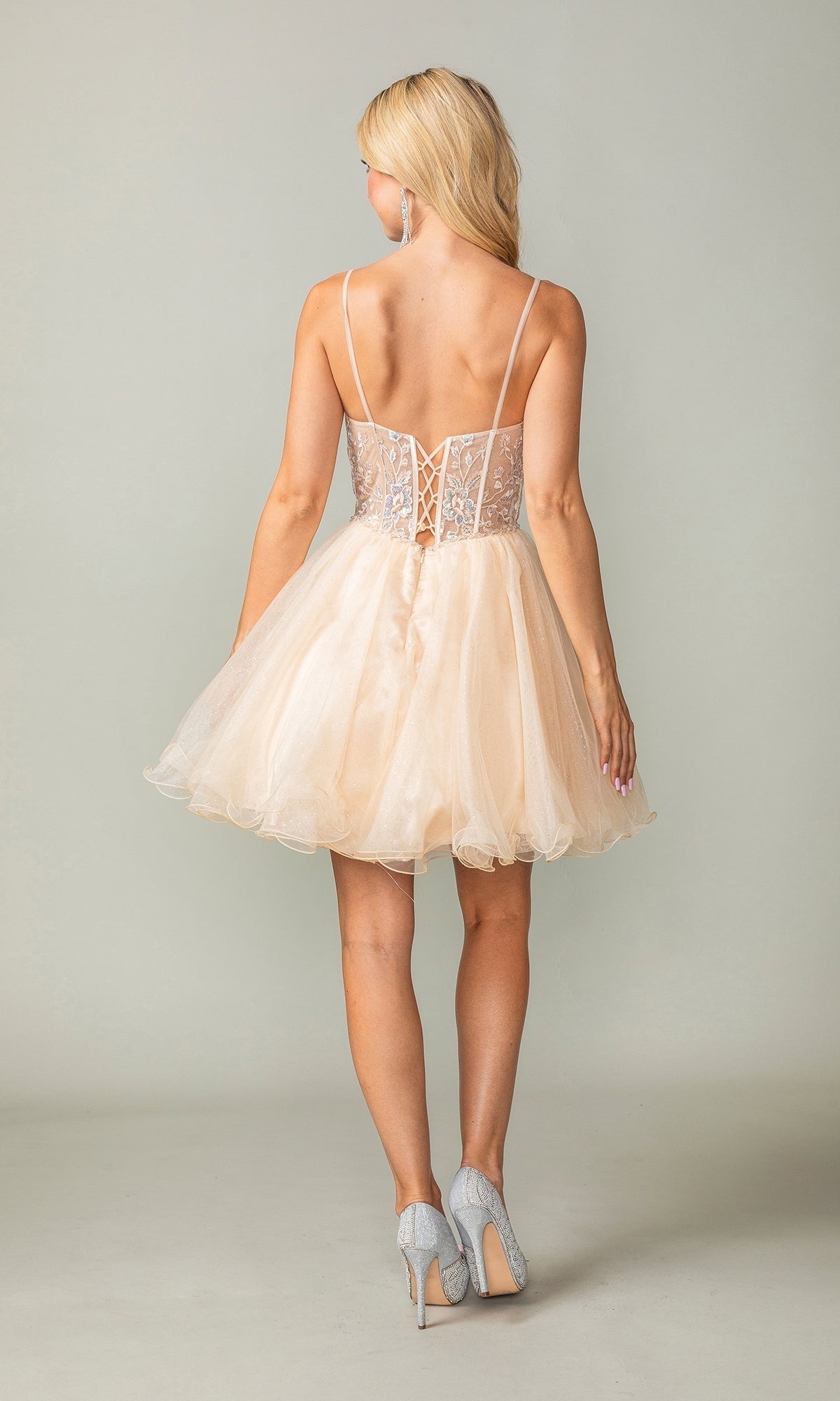 Woman facing away, wearing the Lace-Up Short Babydoll Homecoming Dress 3352 in light beige tulle with a corset-style bodice and thin straps, paired with silver high heels.