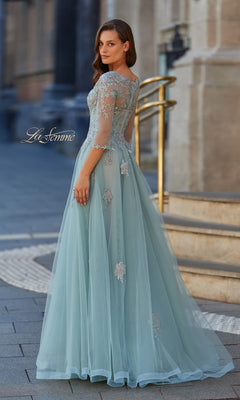 A woman stands outdoors on a paved path, looking over her shoulder in the La Femme 33536 long formal dress—light blue, floor-length tulle with floral embroidery.