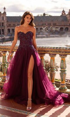 A woman in the Long Prom Dress: La Femme 33551 in Dark Berry, with a strapless beaded lace applique and sheer corset bodice, stands on an ornate balcony against stunning architectural buildings.