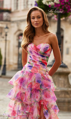 A woman stands outdoors on a city street wearing the La Femme 33555 long prom dress, featuring a strapless sweetheart neckline, tiered ruffles with florals, and a lace-up open back.