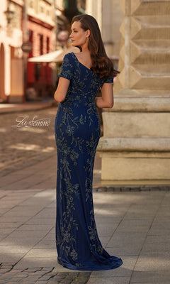 A woman stands outdoors on a cobblestone street, wearing the Long Formal Dress: La Femme 33577—a fitted navy gown with beaded floral embroidery and short sleeves—her back elegantly turned to the camera.
