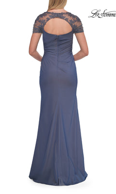 A graceful mother of the bride stands with her hands clasped behind, wearing the Back Cut-Out Long Formal Dress: La Femme 33579—a stunning blue floor-length gown featuring short lace sleeves and an elegant open-back detail.