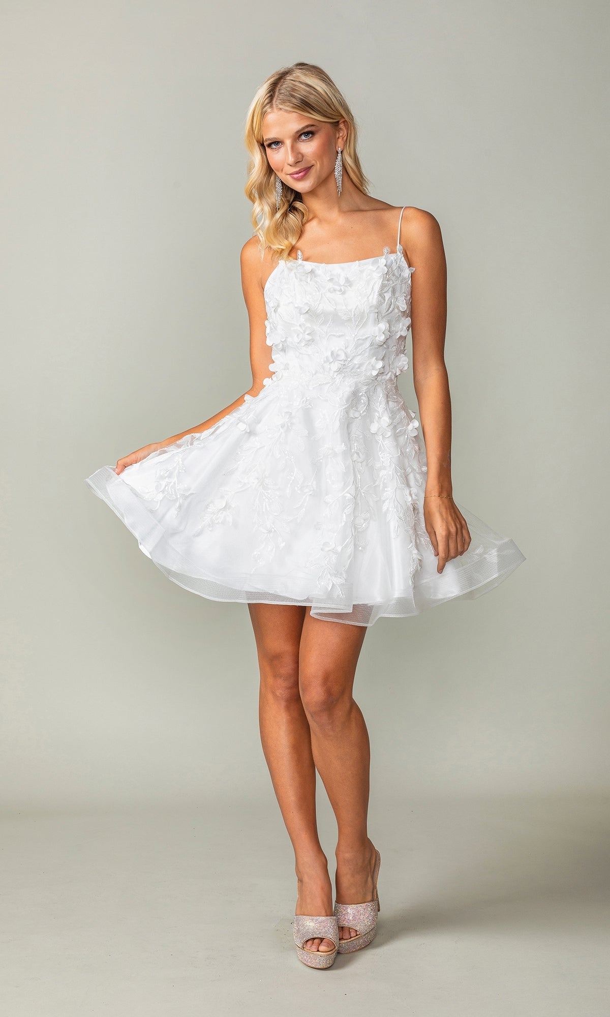 A woman models the Embellished Short Babydoll Homecoming Dress 3357, featuring 3-D floral details and spaghetti straps, as she stands against a plain background holding out her skirt.