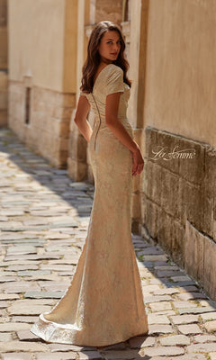 A woman stands on a cobblestone path in the fitted, floor-length La Femme 33598 long formal dress with short sleeves, glancing over her shoulder.