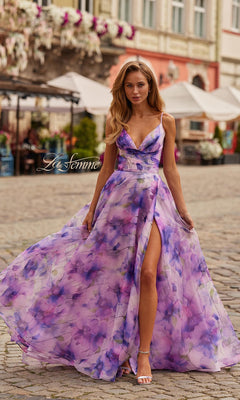 A woman wearing the La Femme 33606 long prom dress in multi stands on a cobblestone street, with outdoor cafes and buildings in the background. The dress features a flowing chiffon fabric, floral print, an a-line skirt, and adjustable spaghetti straps.