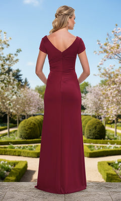 A woman stands with her back to the camera in a formal garden, wearing the Long Prom Dress: La Femme 33616, a floor-length column dress with short sleeves.