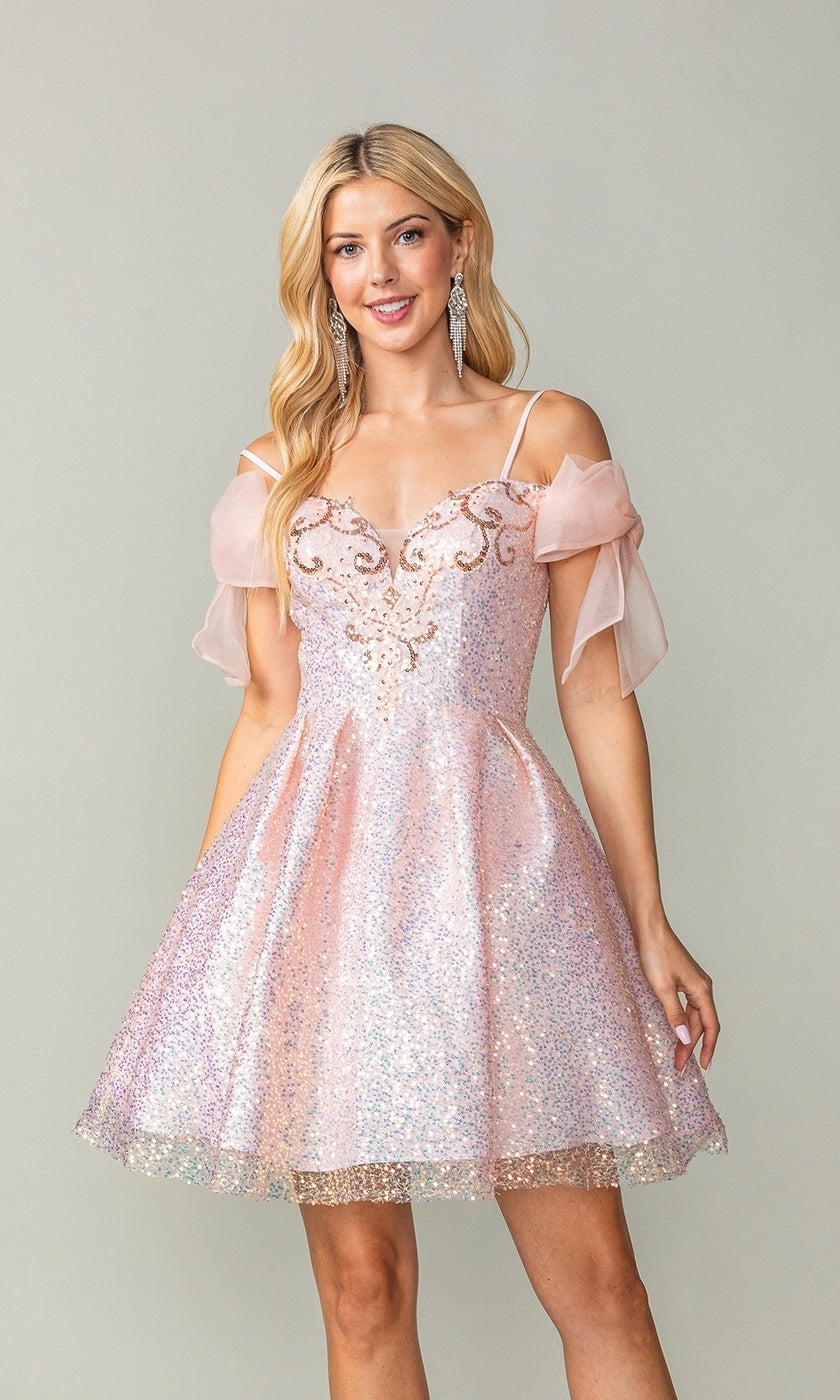 A woman wears the Shoulder-Bow Short Glitter Homecoming Dress 3363 in sparkly pink, featuring spaghetti straps, sheer off-the-shoulder sleeves, intricate bodice details, and a corset lace-up back against a plain background.