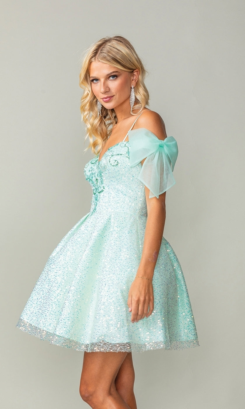 Woman wears the Shoulder-Bow Short Glitter Homecoming Dress 3363—a light blue, off-the-shoulder dress with sheer puff sleeves and a lace-up corset back—standing against a plain light background.