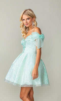 Woman wears the Shoulder-Bow Short Glitter Homecoming Dress 3363—a light blue, off-the-shoulder dress with sheer puff sleeves and a lace-up corset back—standing against a plain light background.