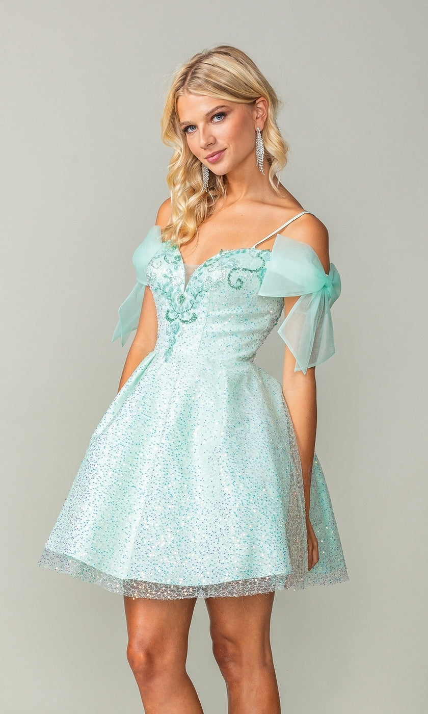 A woman models the Shoulder-Bow Short Glitter Homecoming Dress 3363, featuring a light blue hue, off-the-shoulder style with sheer tulle details, thin straps, and a lace-up corset back against a plain light background.