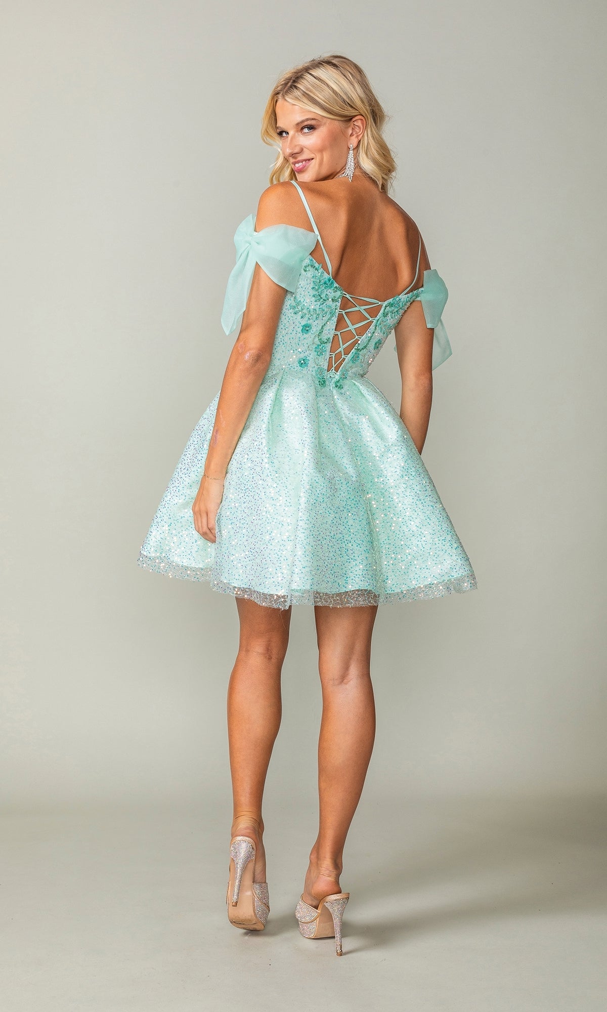 A woman models the Shoulder-Bow Short Glitter Homecoming Dress 3363 in light blue, featuring off-the-shoulder sleeves, a corset lace-up back, and sparkling fabric while looking over her shoulder.