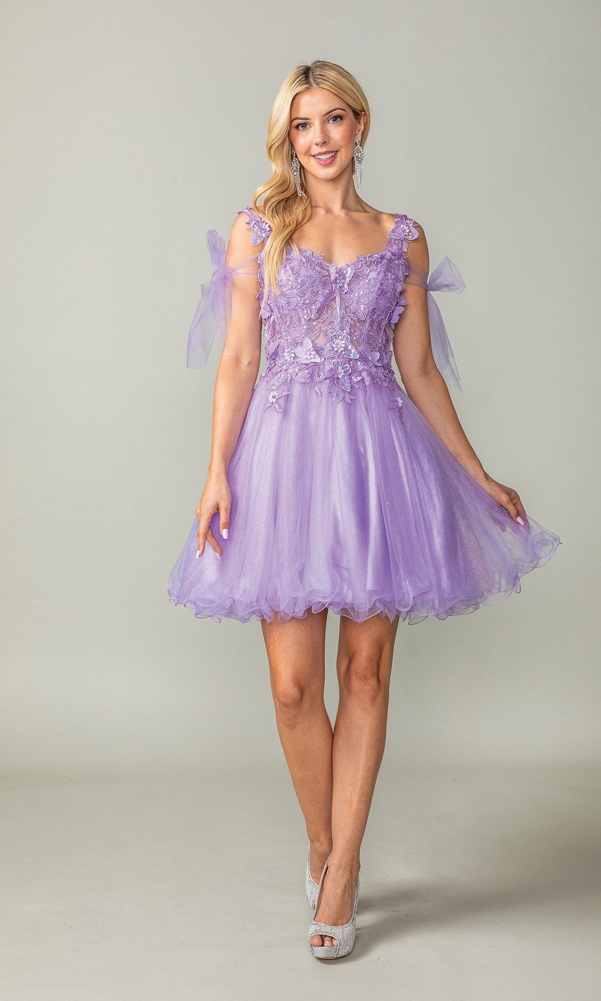 A woman stands against a plain background wearing the Dancing Queen 3365 short homecoming dress in lilac, which features a glitter tulle skirt, floral lace details, and sheer shoulder bows.