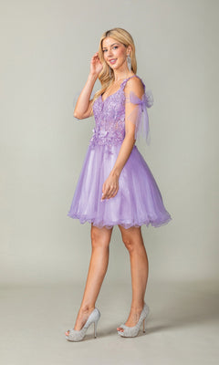 A woman stands and smiles, wearing the Dancing Queen 3365 short homecoming dress—a lavender babydoll-style piece featuring floral appliqué details and a glitter tulle skirt—paired with silver high heels, against a plain light background.