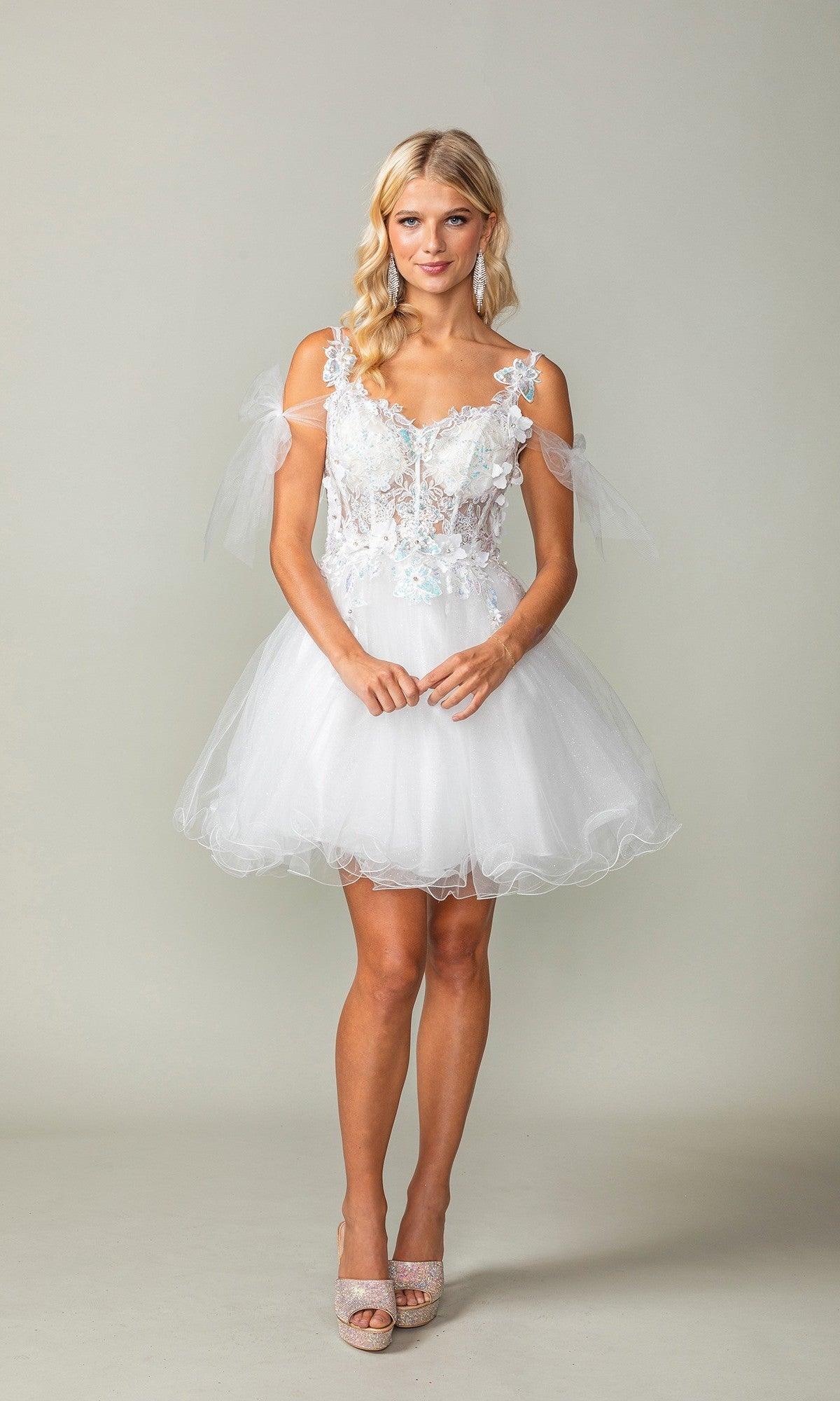 A woman poses against a plain background wearing the Dancing Queen 3365 short homecoming dress in off white, featuring floral appliqué details, sheer shoulder straps, and a delicate glitter tulle skirt.