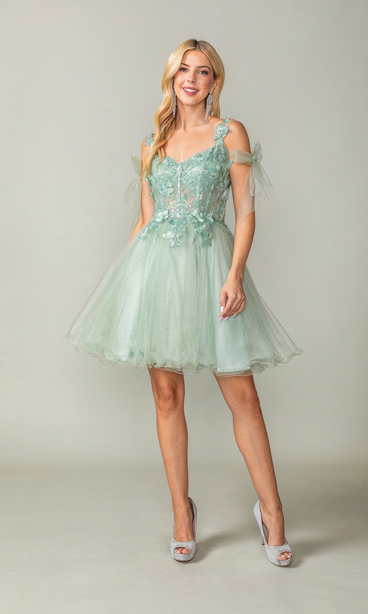 A woman wearing the Dancing Queen 3365 short homecoming dress in sage—a light green, off-shoulder style featuring floral lace details and a glitter tulle skirt—paired with silver peep-toe heels, stands against a plain background.