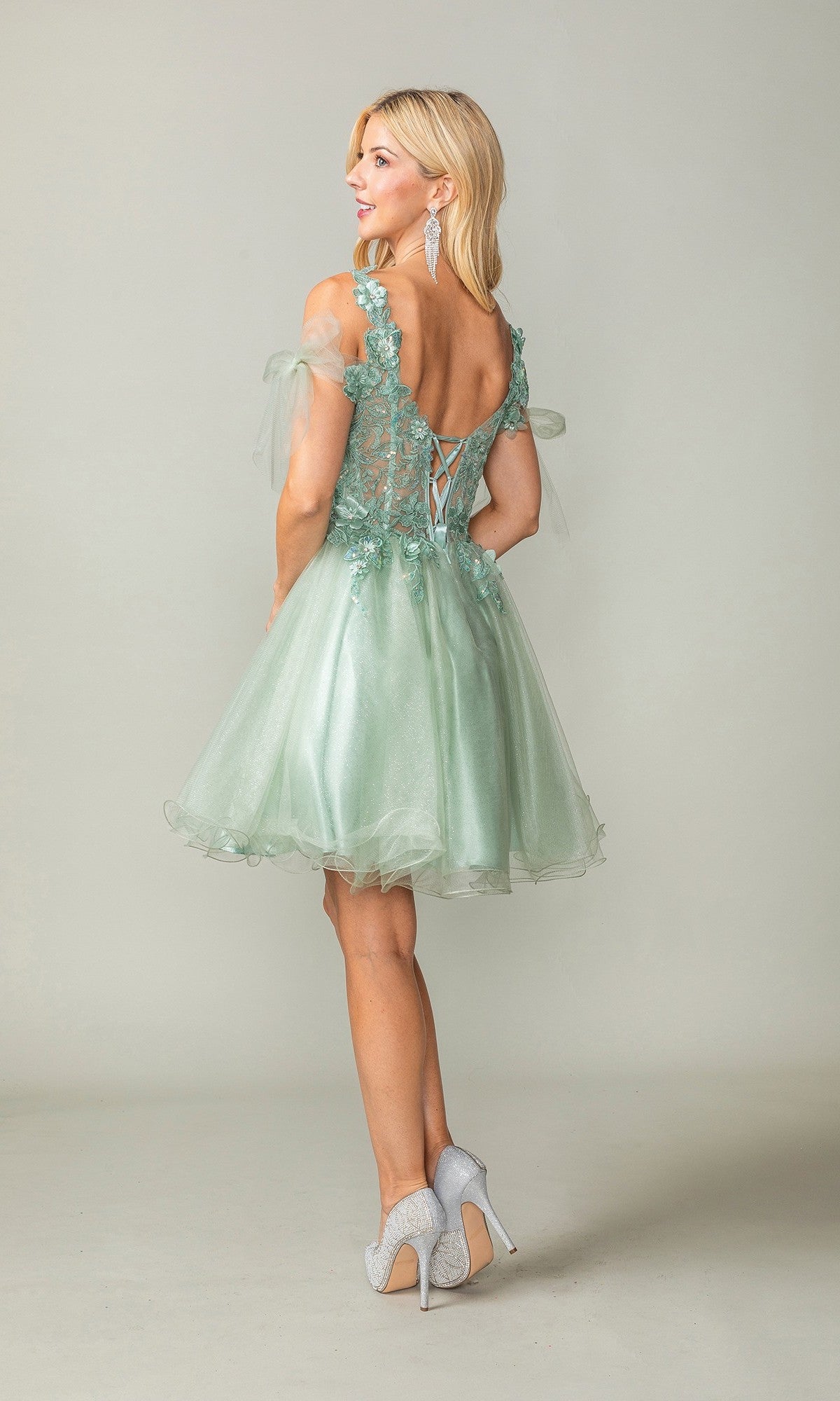 Wearing the Dancing Queen 3365 Short Homecoming Dress, a woman stands on a plain light background. The dress is a light green babydoll style with a lace-up back, sheer sleeves, delicate lace details, and a subtle glitter tulle skirt. She completes her look with gray high heels.