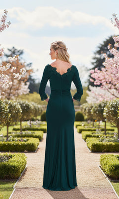 Wearing the La Femme 33658 long prom dress with lace appliqué, a woman stands on a garden pathway, facing away amid blooming trees and manicured hedges.