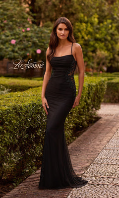 A woman wears the Long Prom Dress: La Femme 33678 in black, featuring lace side panels, while standing on a stone path in a formal garden. The "La Femme" logo is visible on the left side of the image.