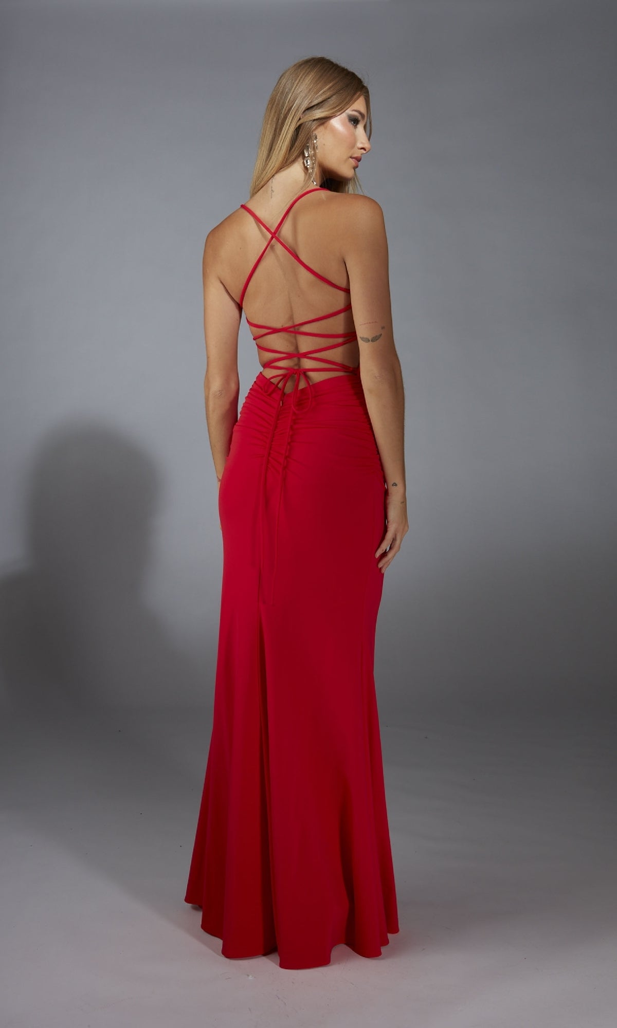 A woman wearing the Aerie Couture 336 long prom dress stands against a plain gray background; the dress features red satin, a fitted bodice, crisscross straps, and a ruched back.