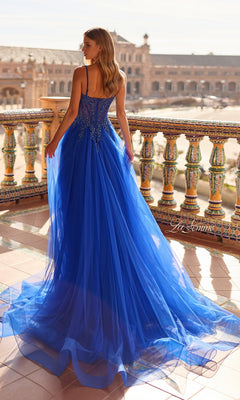A woman wearing the La Femme 33702 long prom dress—an A-line royal blue tulle gown with a beaded lace corset bodice and thin straps—stands on a tiled balcony overlooking a historic plaza.