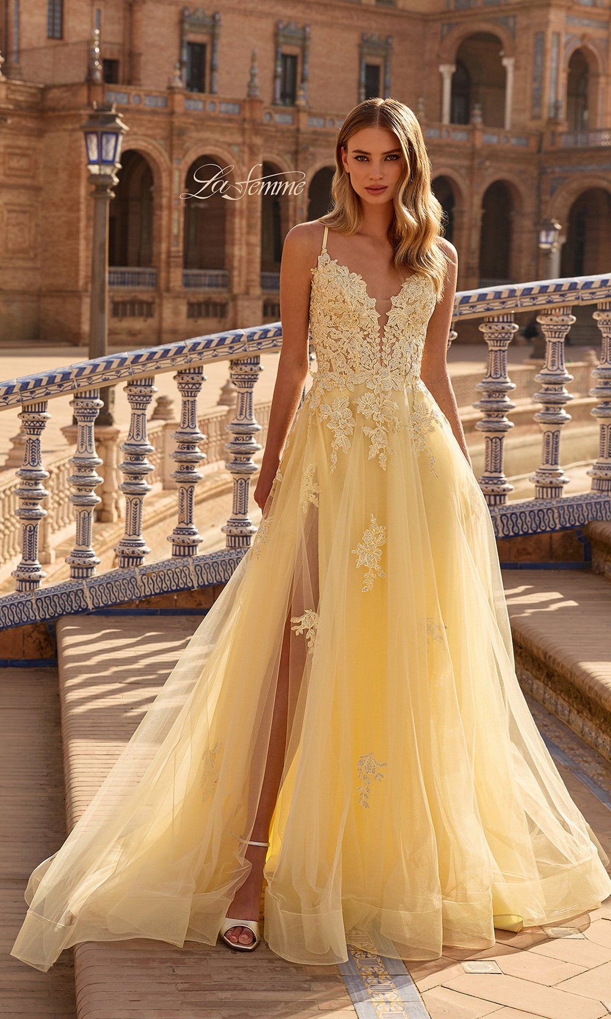 A woman stands outdoors in front of decorative architecture wearing the La Femme 33716 long prom dress—a yellow, sleeveless gown with lace embroidery and a sheer tulle skirt.