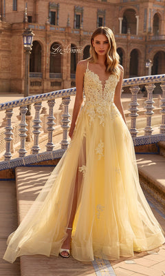A woman stands outdoors in front of decorative architecture wearing the La Femme 33716 long prom dress—a yellow, sleeveless gown with lace embroidery and a sheer tulle skirt.