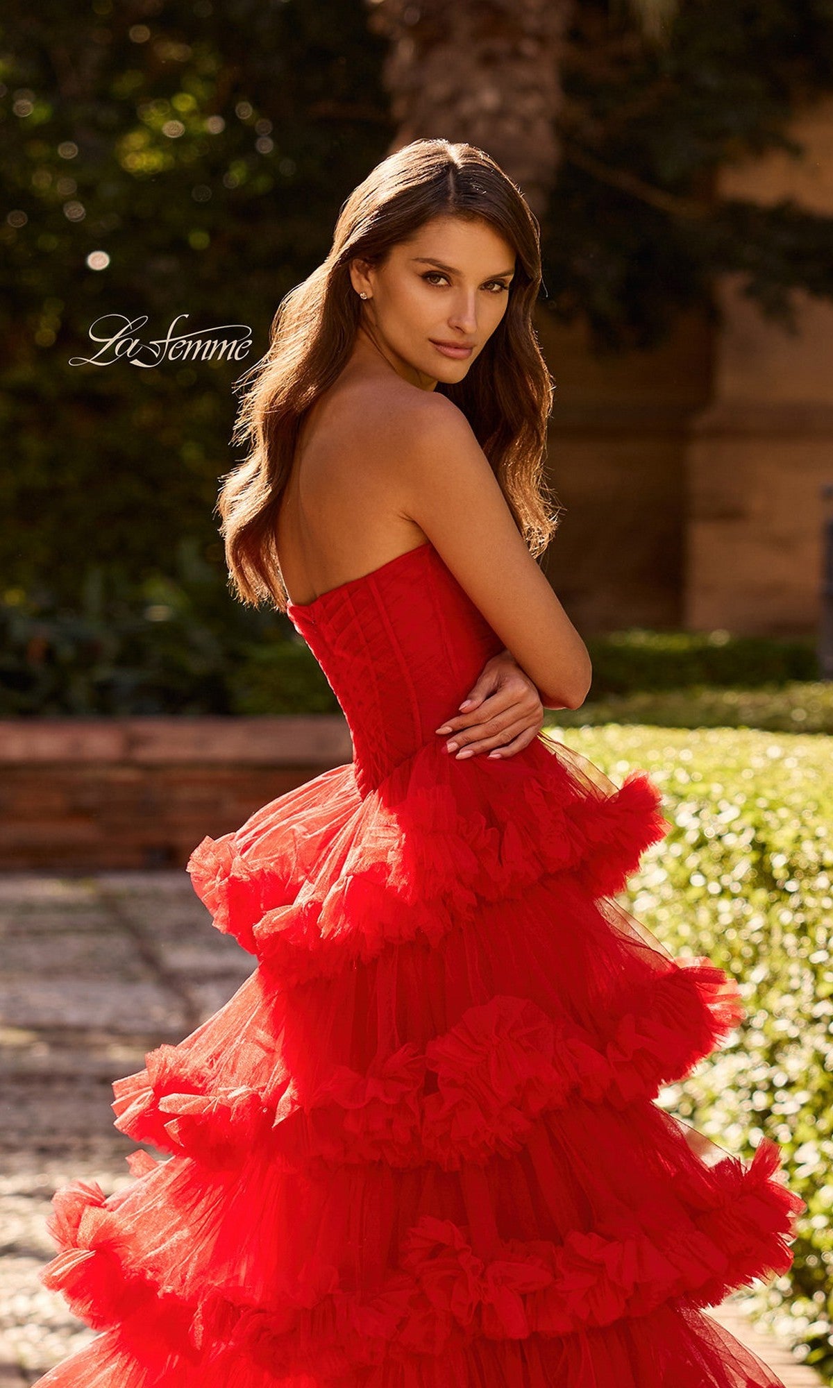 A woman stands outdoors on a stone path in the Long Prom Dress: La Femme 33717, featuring a strapless bodice and tiered ruffle tulle skirt, with greenery behind her. The 