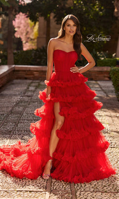 A woman poses outdoors in the La Femme 33717 Long Prom Dress—a strapless ball gown with bold red tiered ruffles and a high slit—standing on a stone path amidst greenery and pink blossoms.