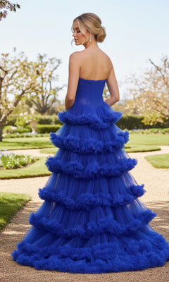 A woman in the La Femme 33717 Strapless Tiered Prom Ball Gown stands on a garden path, her royal blue ball gown flowing behind her amid trees and lush greenery.