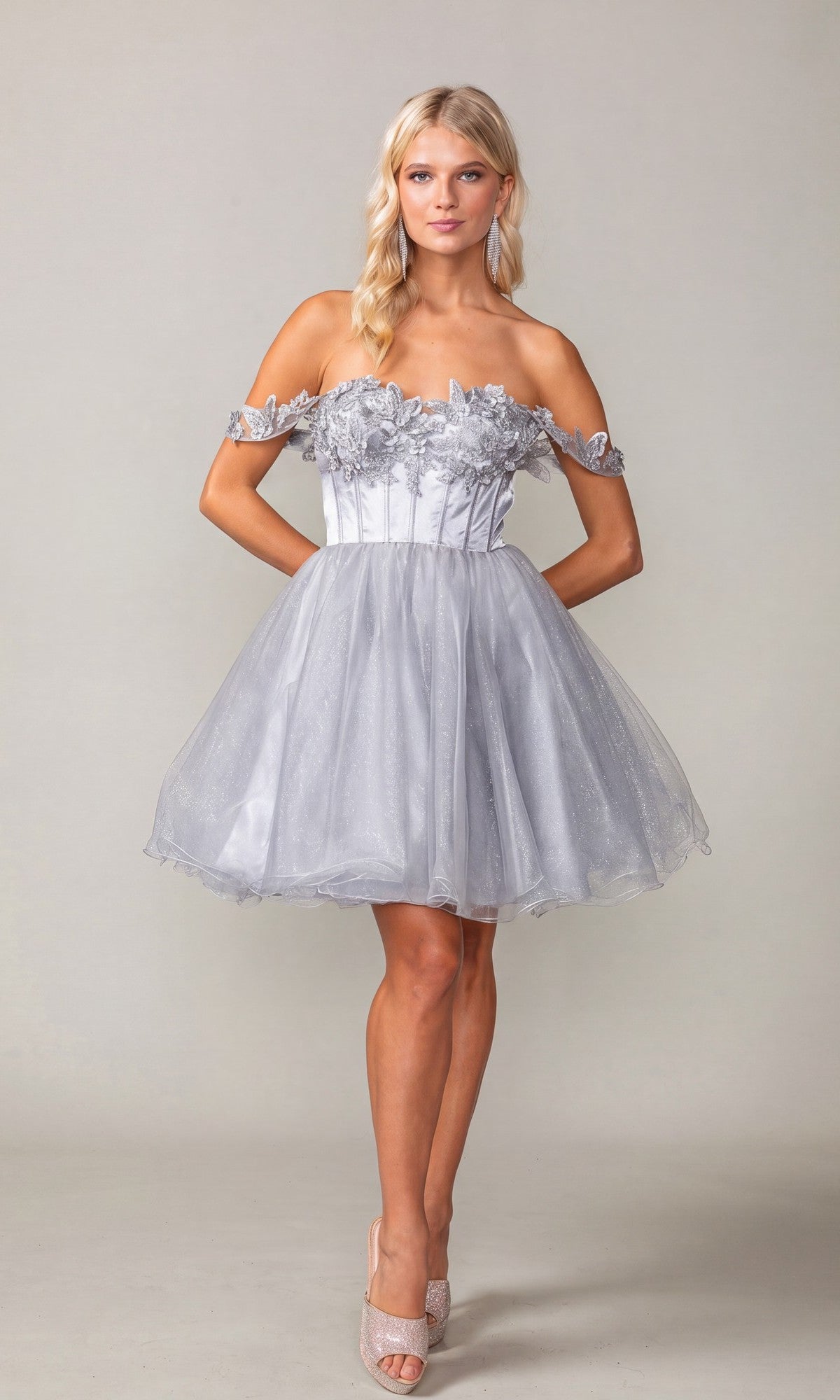 A woman stands with her hands in the pockets of the Lace-Up Corset Mini Hoco Dress: Dancing Queen 3371 in shimmering silver, featuring a short, off-the-shoulder design and delicate floral detailing, against a neutral background.