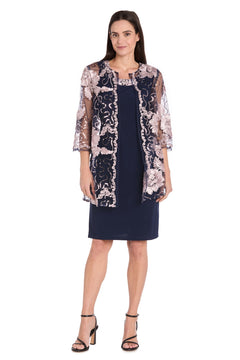 A woman stands facing forward in the R&M Richards 3371 Short Navy Jacket Party Dress, featuring a navy cocktail dress with a sheer floral embroidered jacket and black heeled sandals—an elegant semi-formal look for parties.