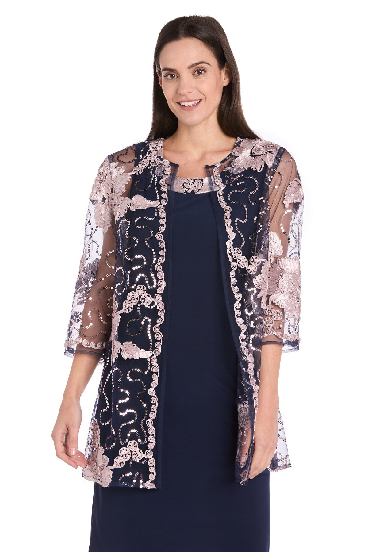 A woman stands smiling at the camera in the R&M Richards 3371 Short Navy Jacket Party Dress, featuring a sheer floral embroidered overlay and three-quarter sleeves—an elegant, charming semi-formal look for any occasion.