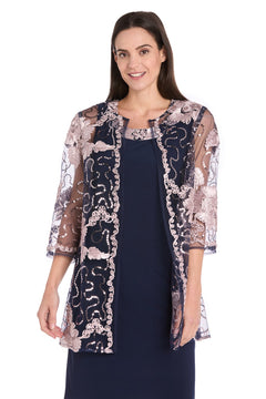 A woman stands smiling at the camera in the R&M Richards 3371 Short Navy Jacket Party Dress, featuring a sheer floral embroidered overlay and three-quarter sleeves—an elegant, charming semi-formal look for any occasion.