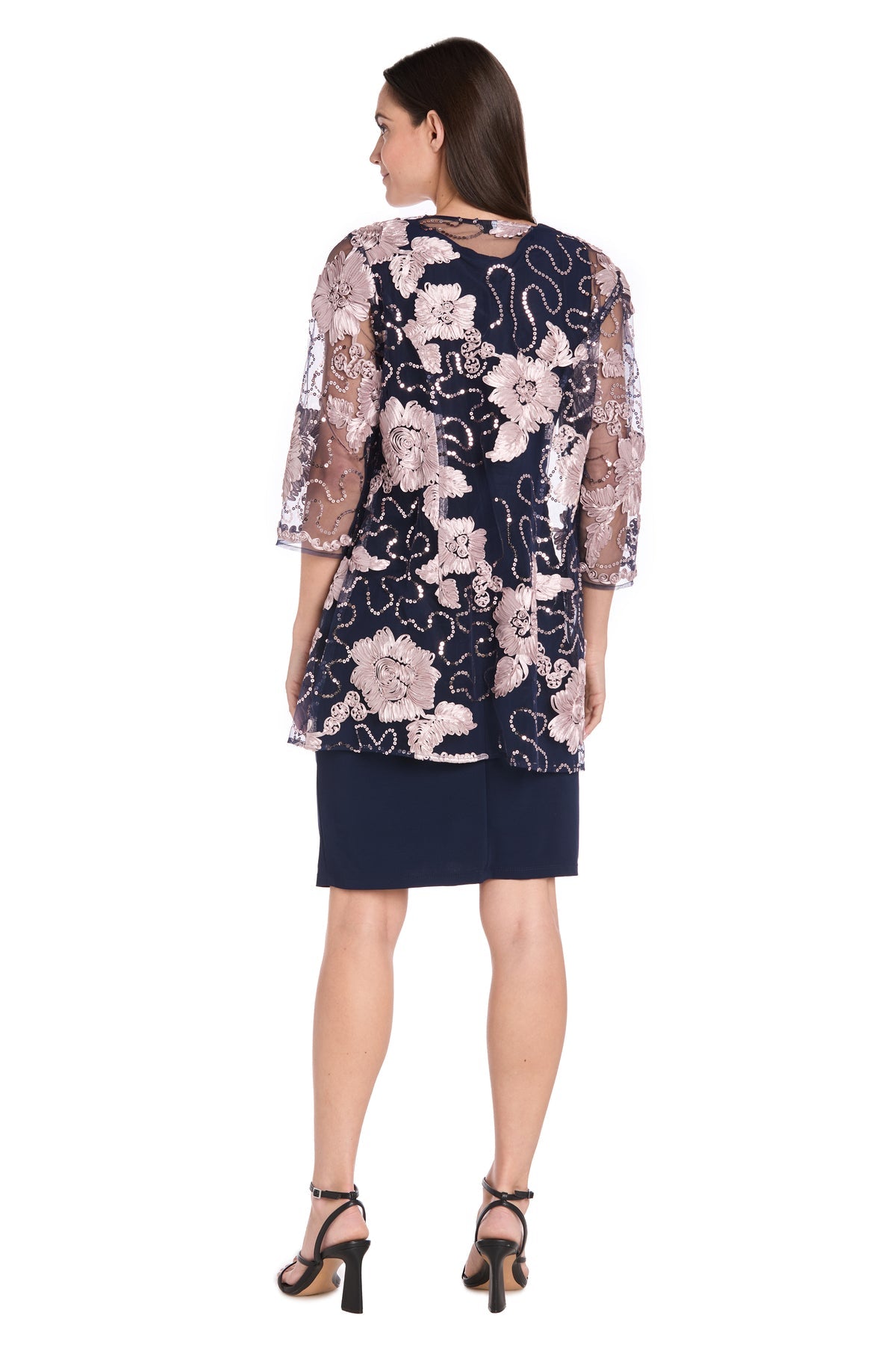 A woman, back to the camera, wears the R&M Richards 3371 Short Navy Jacket Party Dress with a sheer floral overlay and black heeled sandals.