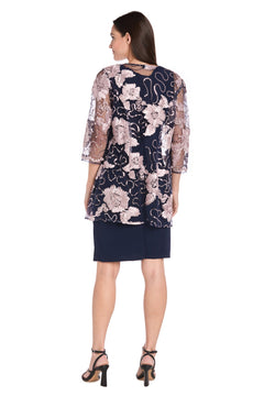 A woman, back to the camera, wears the R&M Richards 3371 Short Navy Jacket Party Dress with a sheer floral overlay and black heeled sandals.