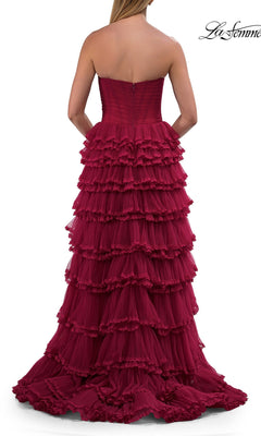 A woman stands facing away from the camera, wearing the La Femme 33727 long prom dress—a strapless, floor-length burgundy gown featuring a corset bodice and multiple layered ruffles.