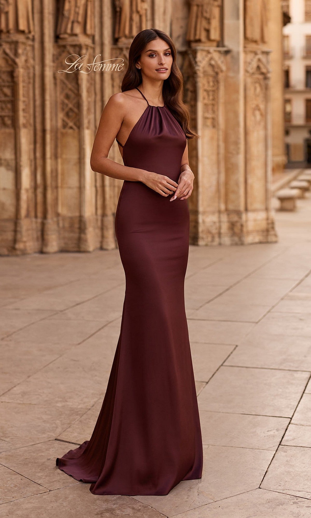 A woman in the La Femme 33732 long burgundy satin prom dress with an open back stands outdoors before ornate stone architecture.