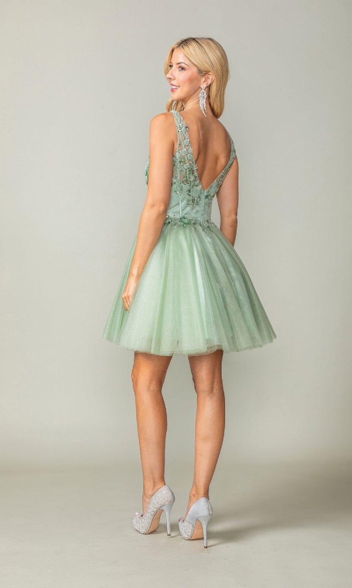 A woman faces away, wearing the Dancing Queen 3374 open-back tulle homecoming dress, a sleeveless, light green knee-length style with floral lace details and high-heeled shoes.