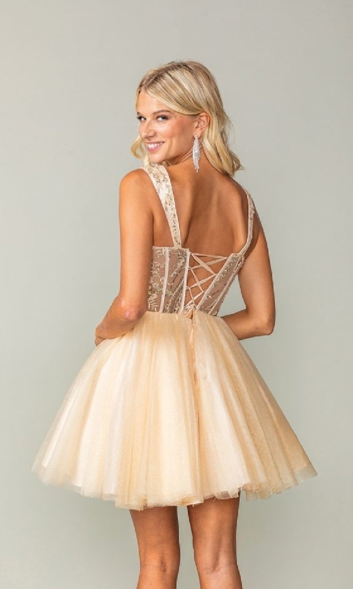 Woman wearing the Lace-Up Open-Back Hoco Dress: Dancing Queen 3375, a short sleeveless light beige tulle dress with flouncy skirt and lace-up back detail, standing and smiling at the camera.
