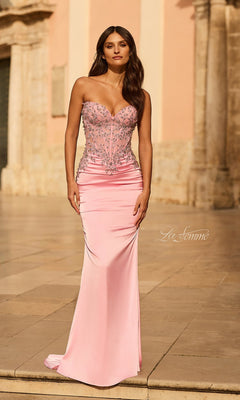 A woman wears the Long Prom Dress: La Femme 33765 in baby pink, featuring a strapless embellished corset bodice with silver details, as she stands outdoors on stone pavement before a historic building.