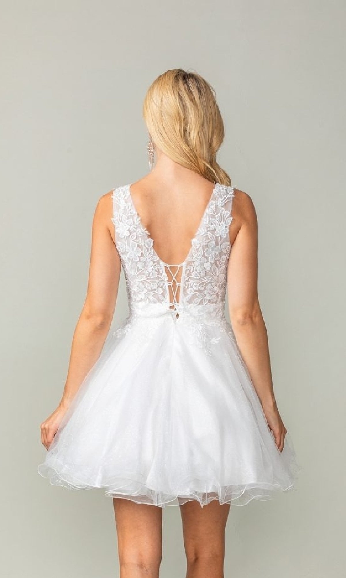 A woman faces away from the camera, wearing the Dancing Queen 3377 Tulle Babydoll Homecoming Dress—a white sleeveless dress with a lace back and V-neckline—against a plain background.