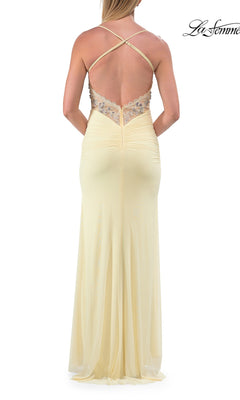 Wearing the Sheer-Corset Long Prom Dress by La Femme 33783, a woman stands facing away from the camera; the gown is long and pale yellow, featuring a low-cut open back with crisscross straps and delicate floral embroidery.