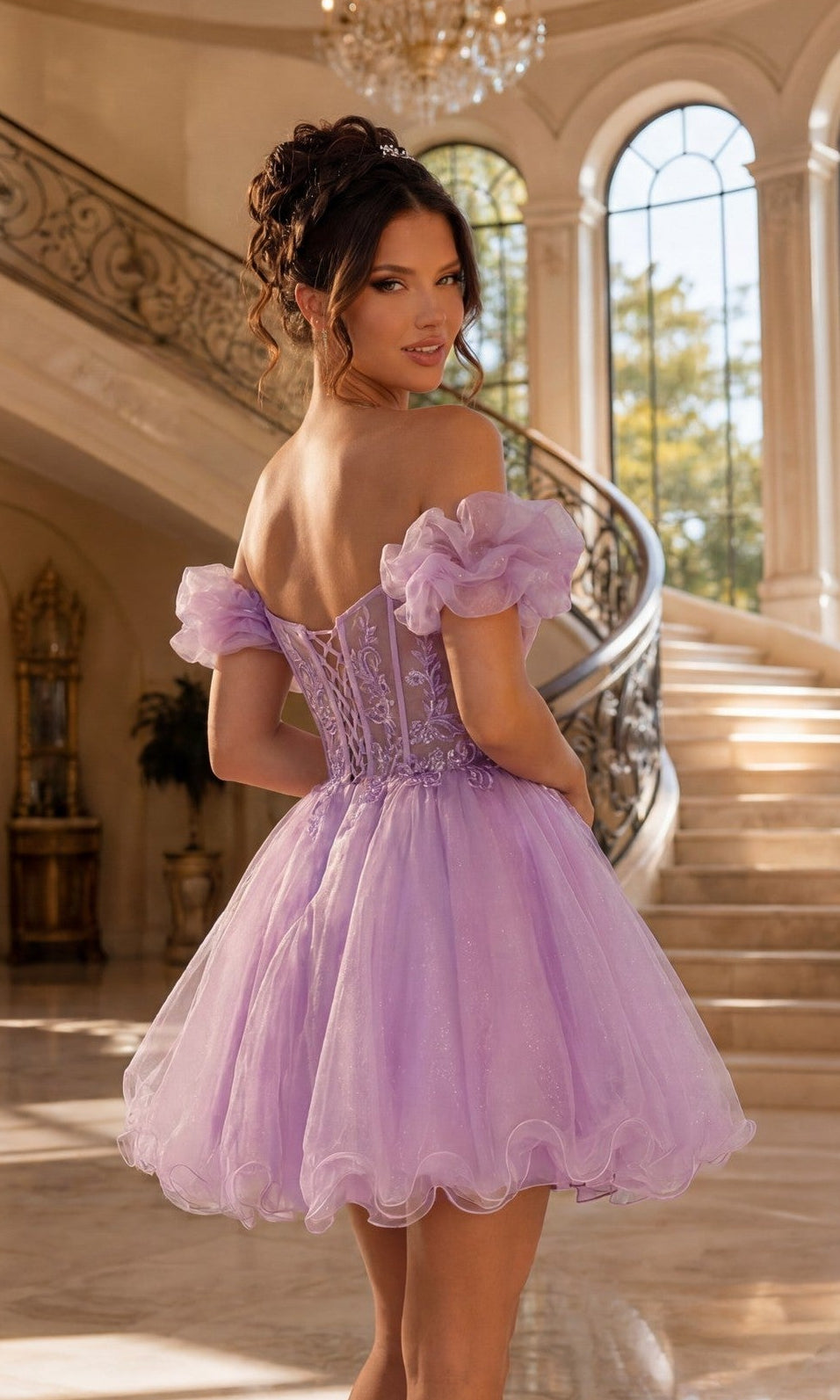 A woman stands indoors in front of a curved staircase and large windows, wearing the Ruffle-Sleeve Short Corset Homecoming Dress 3379, featuring puff sleeves, an off-the-shoulder neckline, and a fitted bodice.